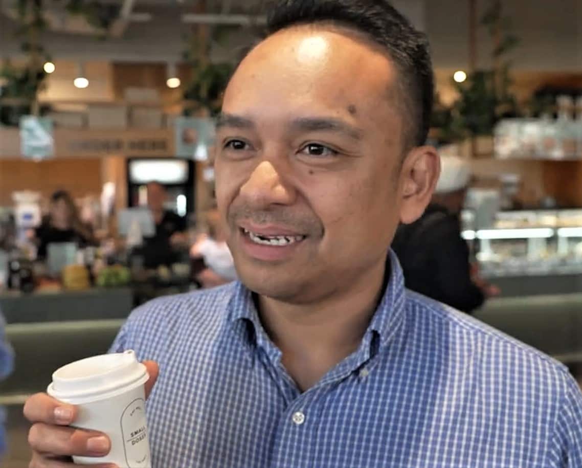 A man in a checked blue shirt stands in a cafe holding a take away coffee cup.