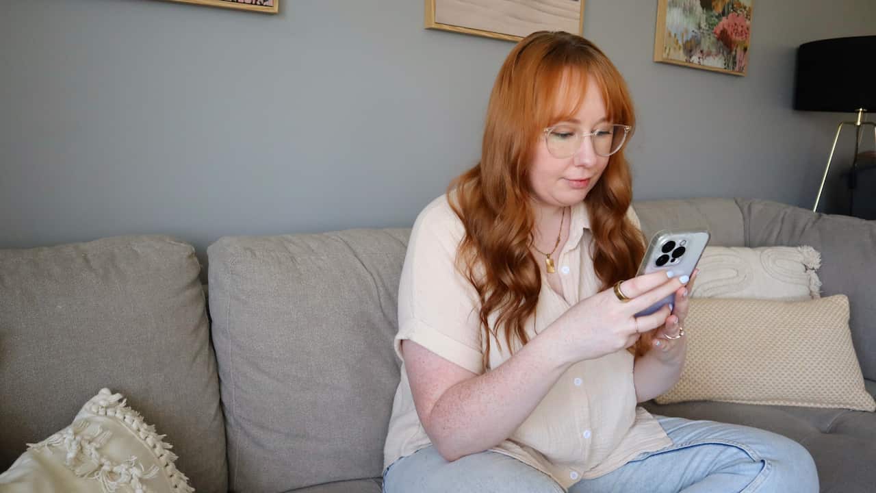 a young woman with red hair sitting cross legged on a couch scrolling on her phone