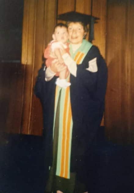 An elderly woman stands in a graduation gown and cap, wearing an Aboriginal flag cape, holding her child, and smiling.
