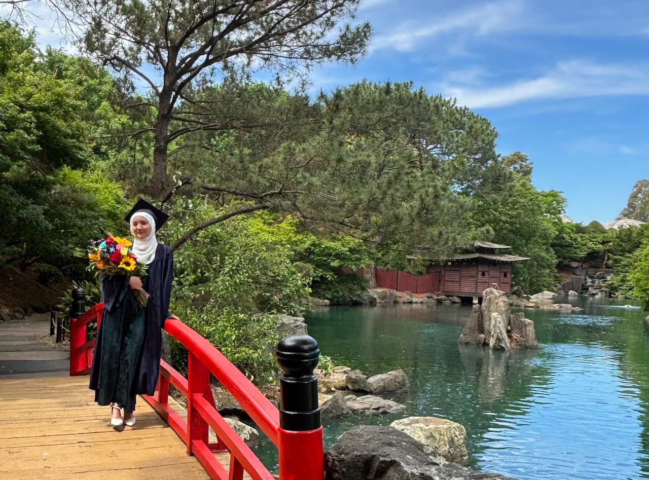 A young Muslim girl in a graduation gown and mortarboard stands near a lake 
