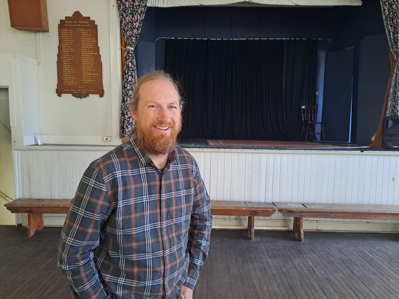 A man in a long-sleeve chequered shirt stands in front of a stage