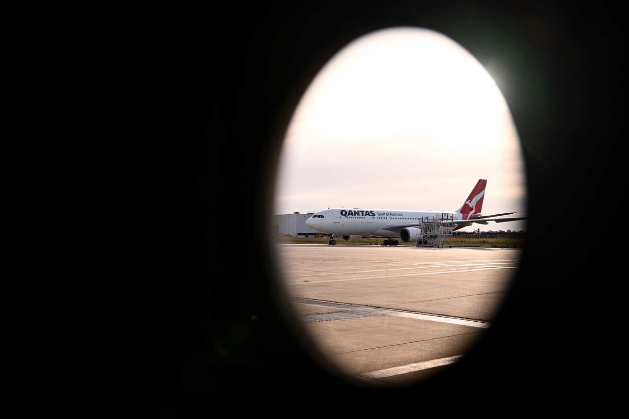 A Qantas aeroplane in focus on the tarmac, seen through a plane window, with a dark oval shaped shadow made by the window framing the scene.