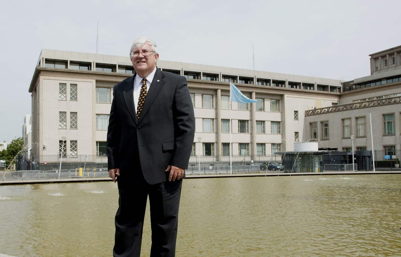 A middle-aged man with white hair and glasses stands outside an official-looking building. He wears a black suit and a brown tie with small spots. In the background, a blue UN flag waves in the wind above a water feature at the building's entrance.