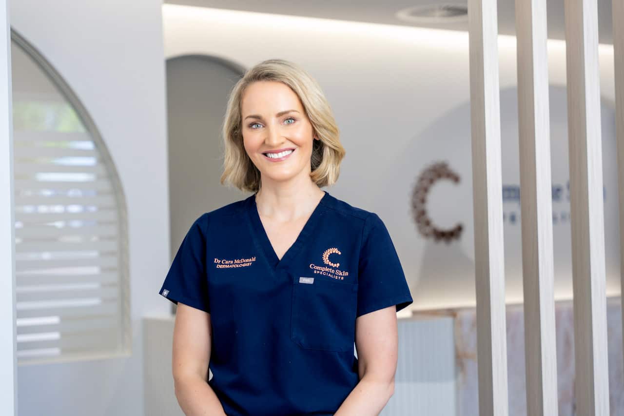 A woman with short blonde hair and wearing a dark blue clinical top smiling in a clinic.