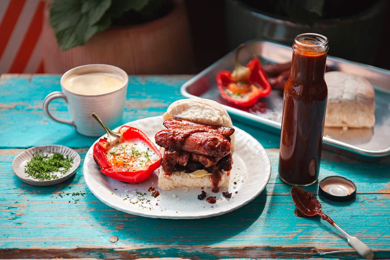 A filled bread roll sits on a white plate, alongside a half capsicum with a cooked egg in it. A cup of coffee sits behind the plate. A tall bottle of brown sauce sits beside the plate. Behind, sitting on the same blue wooden tabletop, is a tray with another bread roll, with fillings and a capsicum half sitting beside it. 