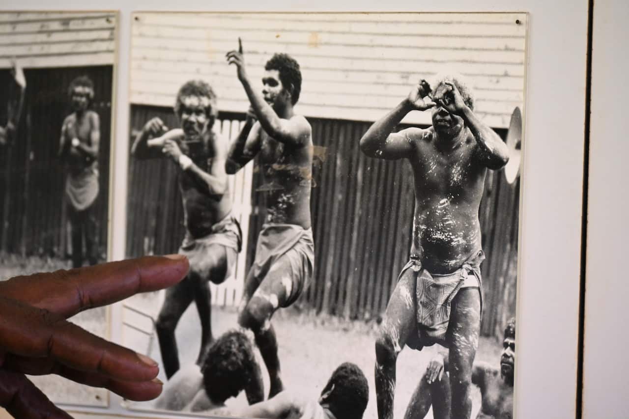 Bernard Tipiloura’s fingers point to an old photograph where his brother (on the right) dances the war dance with two other Tiwi men. They wear traditional paint.