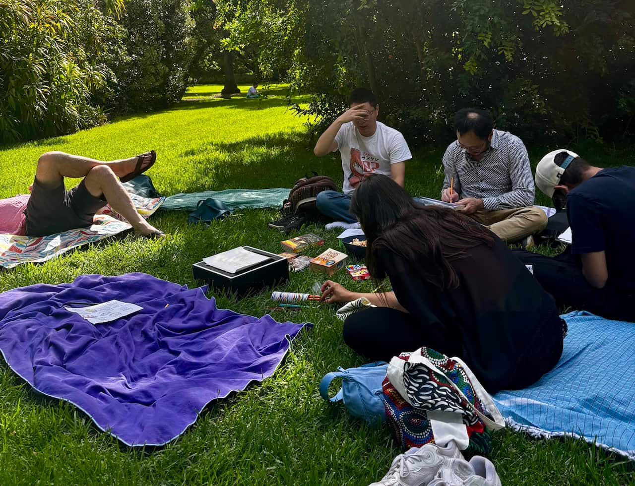 A group of people sitting outside on rugs on the grass together reading or writing.