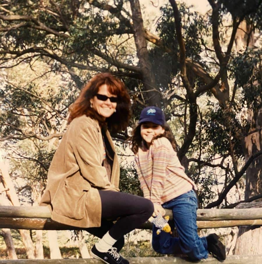 A young girl and her mum sit on a bench. They are looking at the camera and smiling.