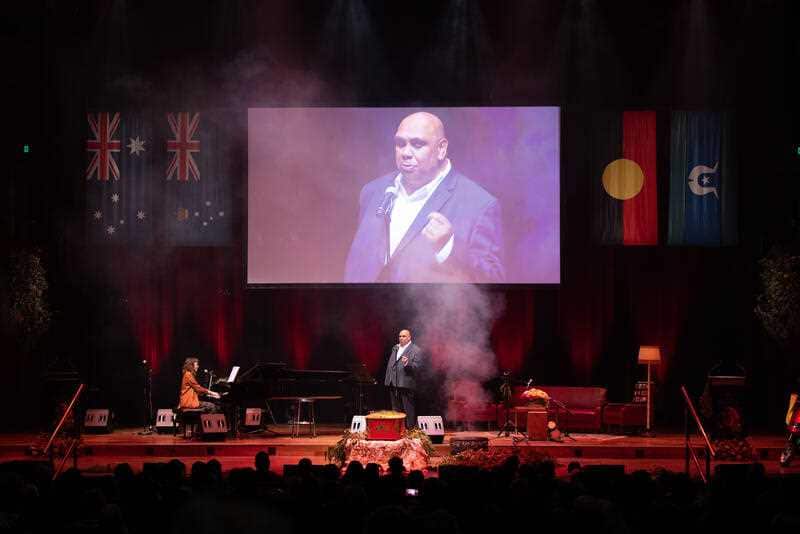 Kutcha edwards performs on stage with his own image superimposed on the projector screen overhead. Smoke wafts across the stage and uncle jack's red and yellow coffin sits brightly lit in centre stage