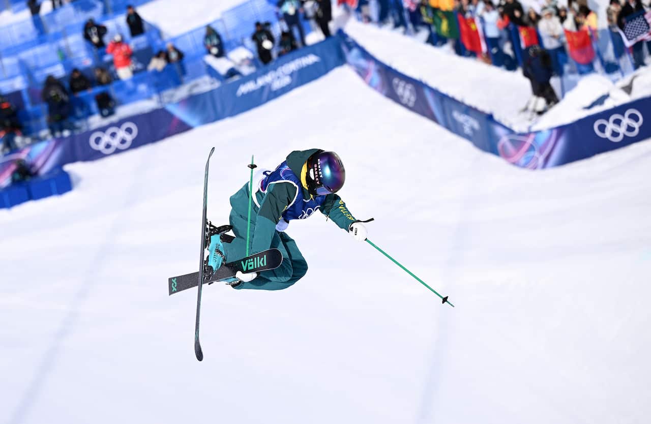 A skier in a green and gold Australian uniform is airborne with her skis crossed. The background is snow with a Winter Olympics banner in the very back.