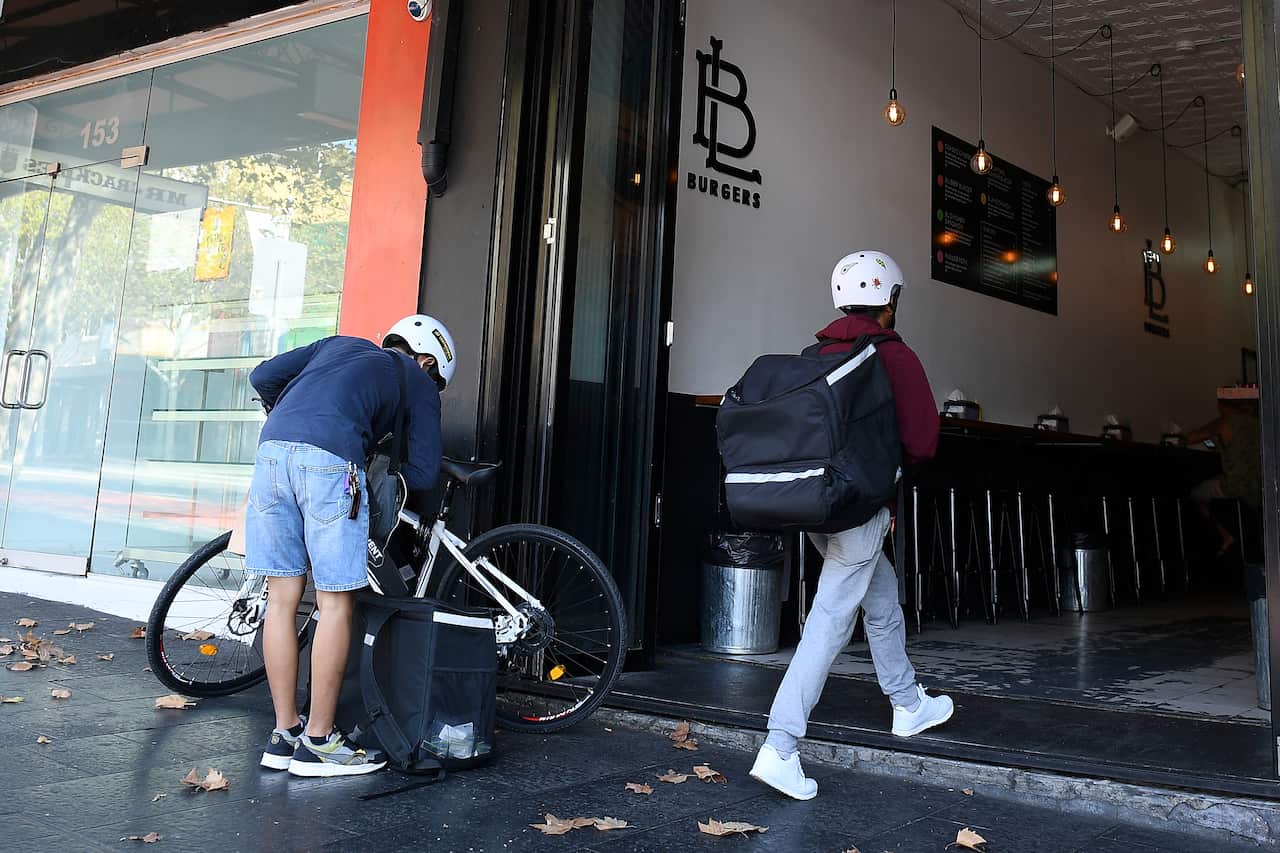 A person walking into a restaurant with an insulated bag on their back as another locks his bike. 