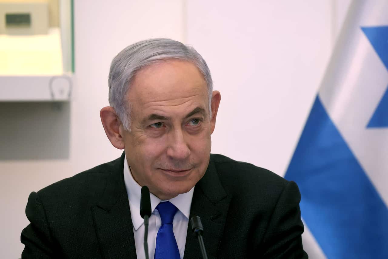 Benjamin Netanyahu sitting behind a microphone. An Israeli flag is behind him.