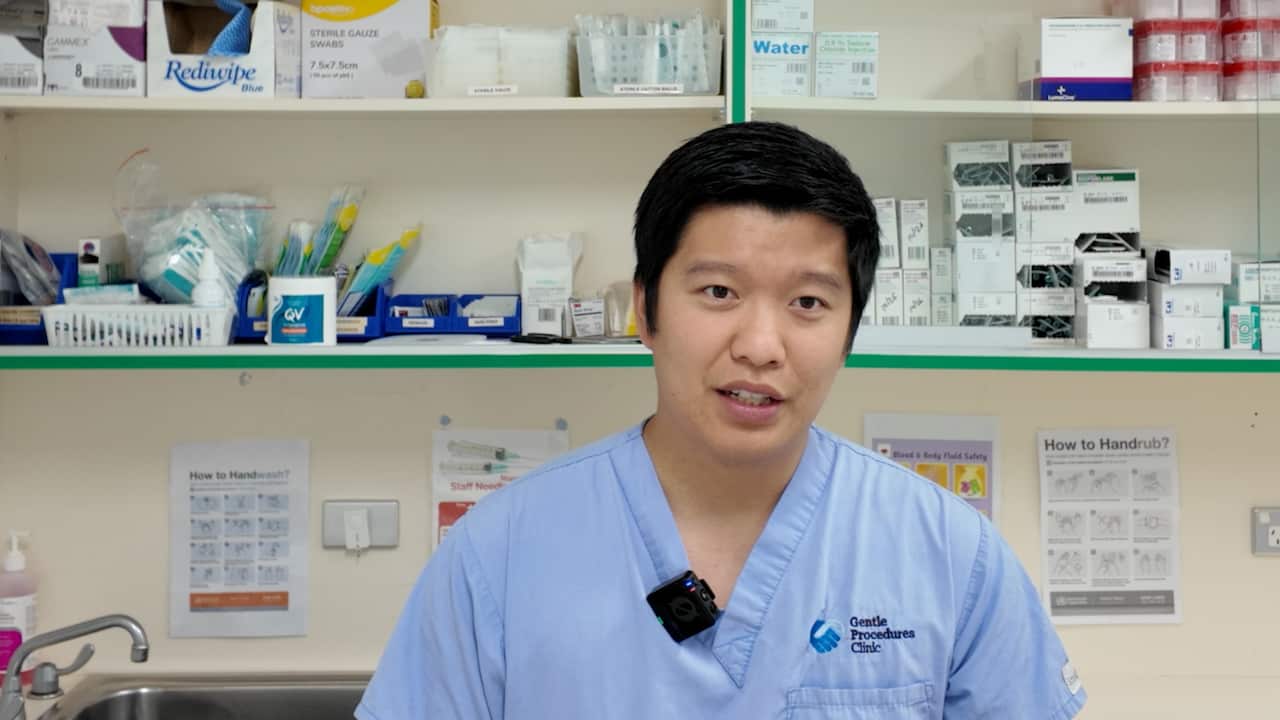 A doctor in front of a clinic room wearing blue scrubs with a tag that reads gentle procedures clinic has a microphone attached to him 