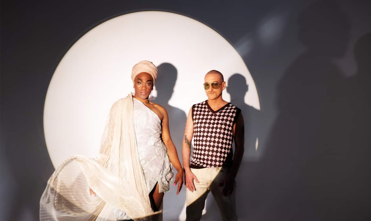 A man wearing an ivory-coloured dress and a cream headwear with a long drape. He is standing next to a man wearing a checked vest and white pants. They are in a spotlight against a black wall.