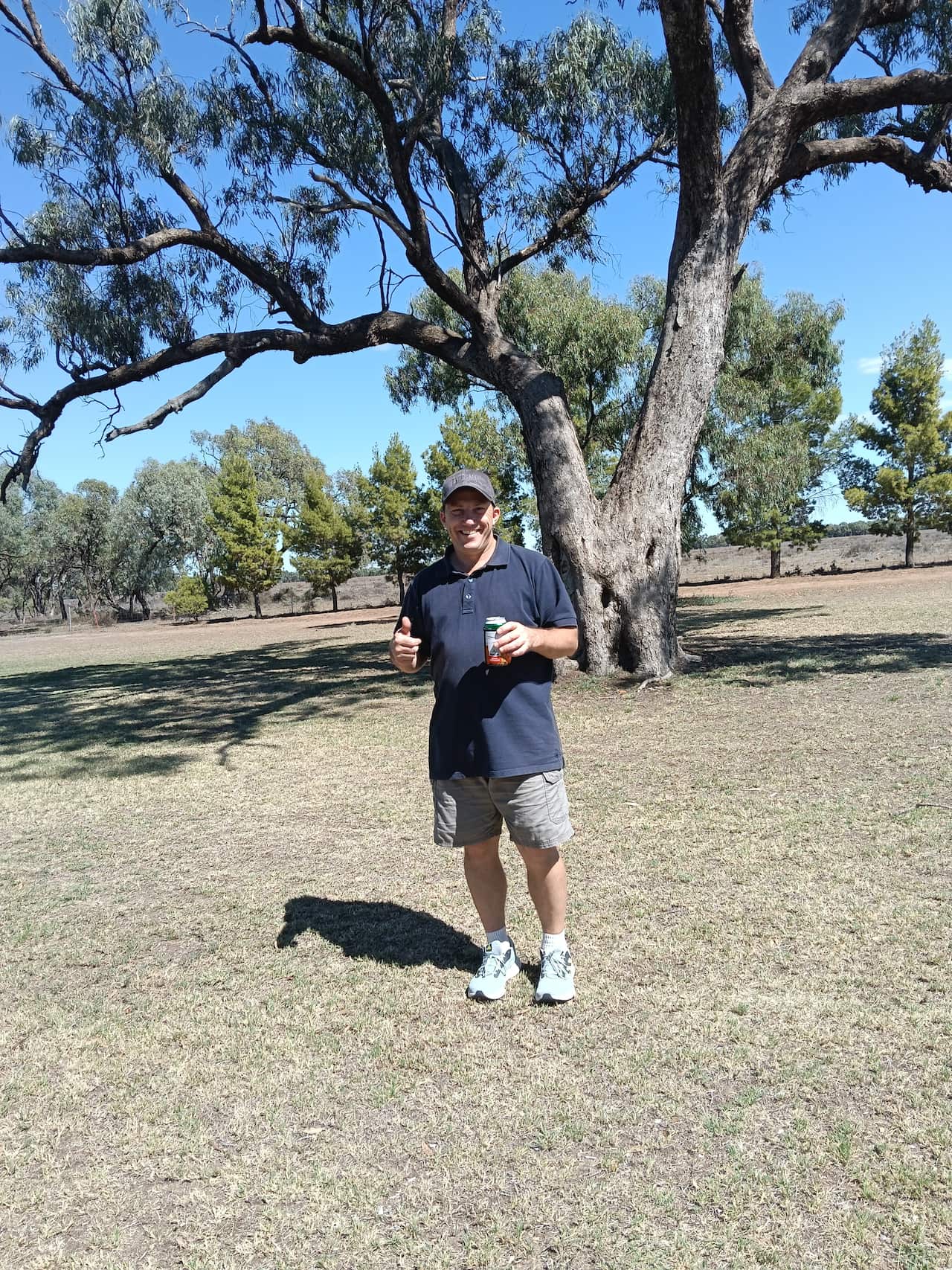 A man standing underneath a tree holds a can and gives a thumbs up 