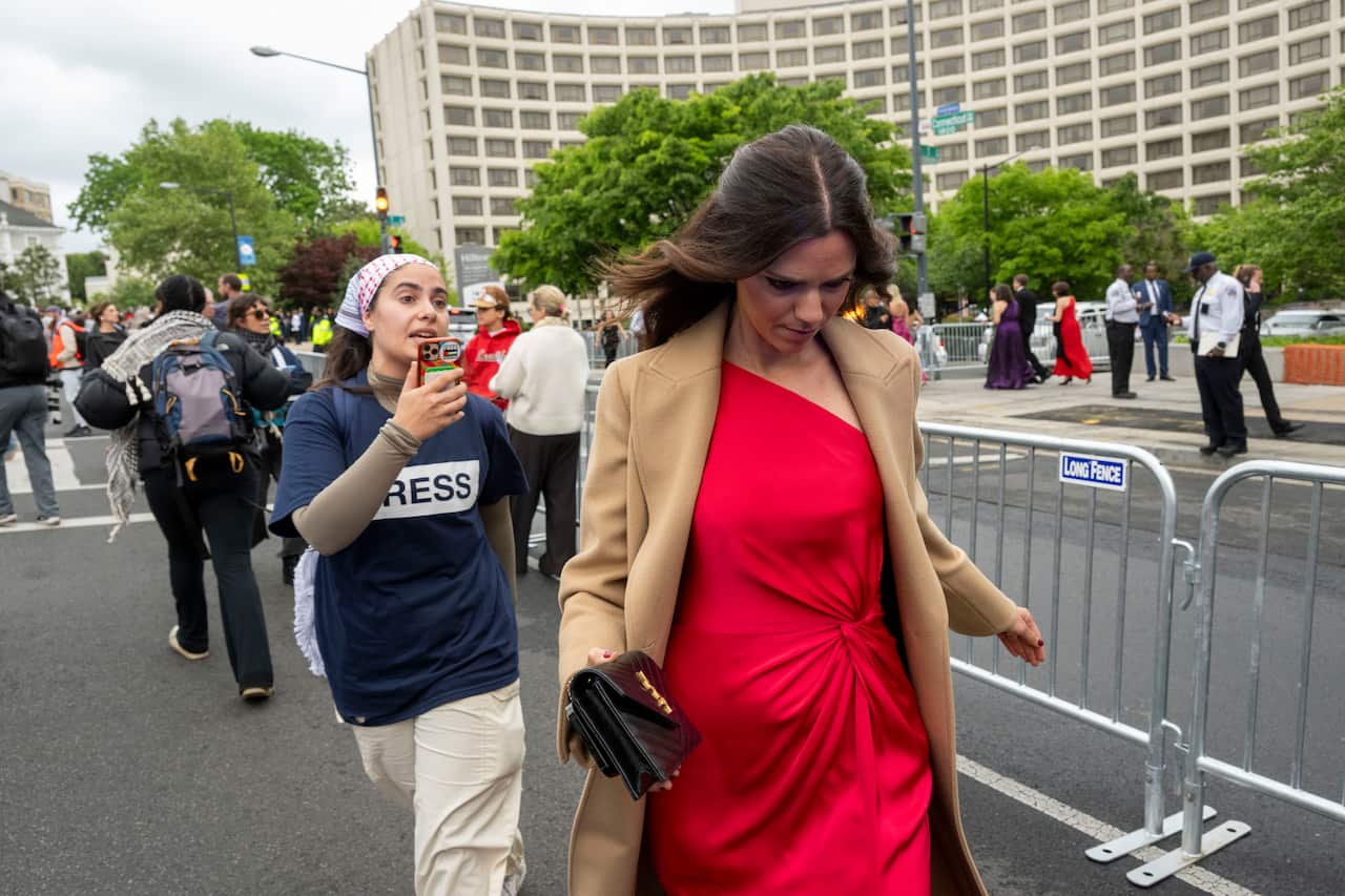A pro-Palestinian protester walks behind a woman in a red gown and tan coat