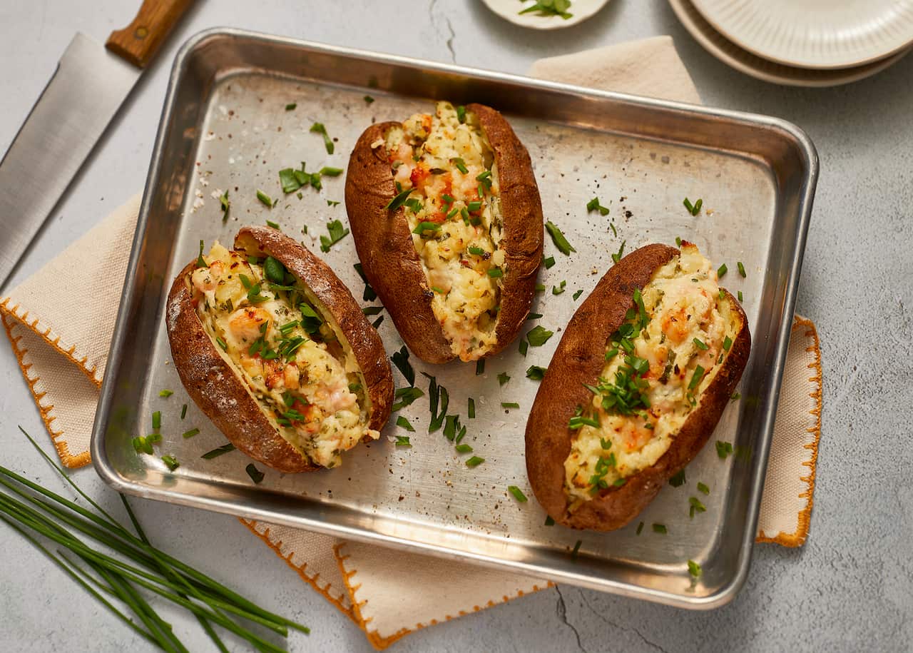 Three baked potatos with a potato and prawn filling sit on a metal baking tray. The potato is scattered with fresh chopped herbs.