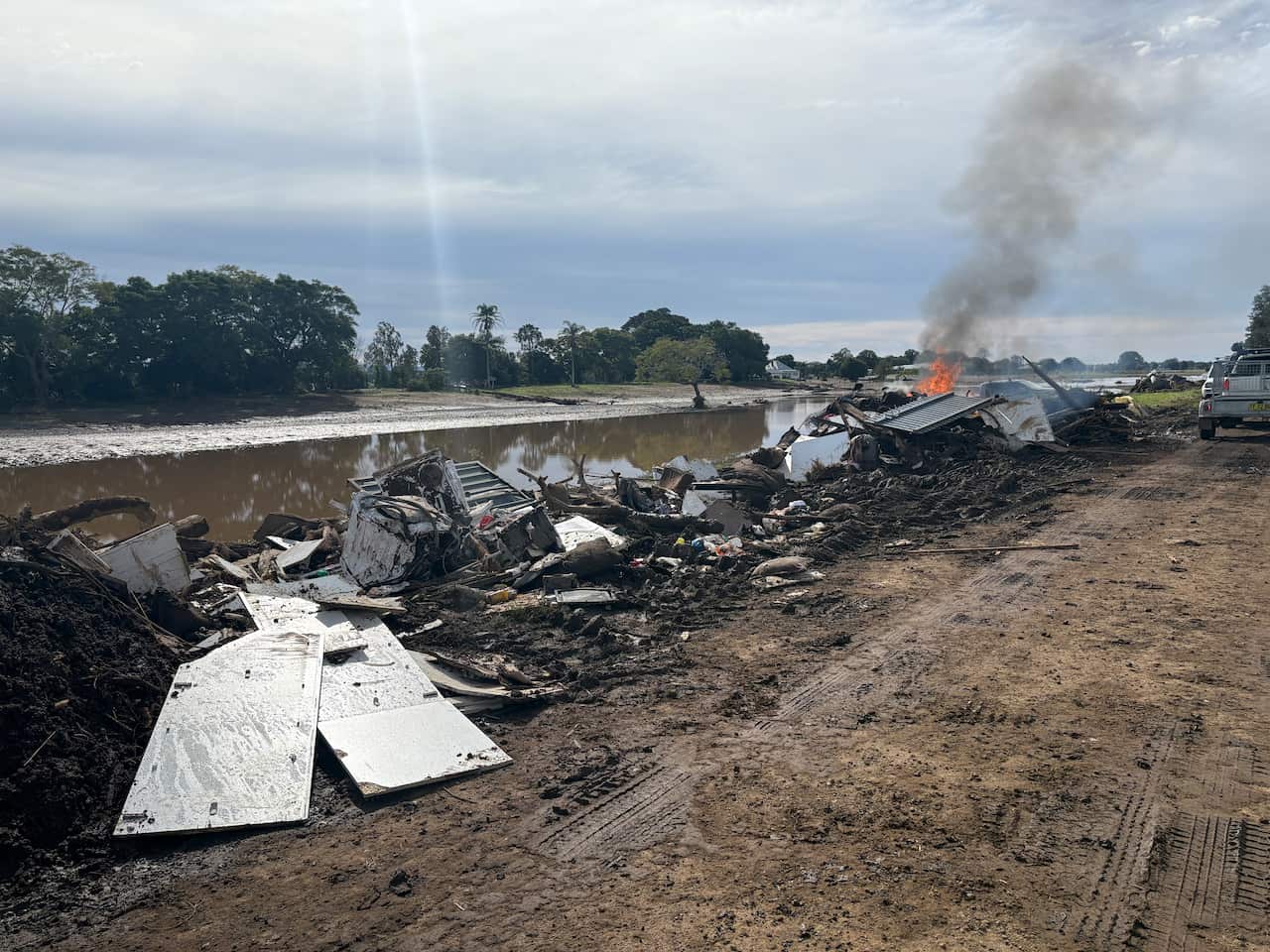 A dirt street lined with miscellaneous rubbish and debris, covered in mud, with a fire burning in the distance. Floodwater is next to the street, with a line of trees in the distance.