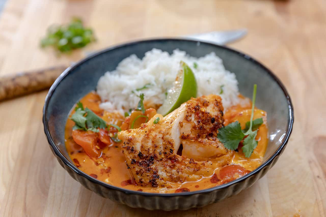 A ineresting pottery bowl holds a soupy-stew with a piece of cooked white fish in an orange-ish creamy sauce, alongside white rice. The bowl is topped with a wedge of lime. 