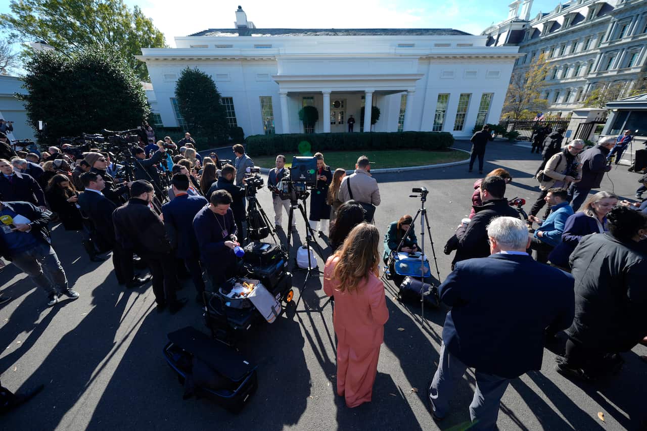 Journalists stand outside a white building, some with cameras and tripod stands.