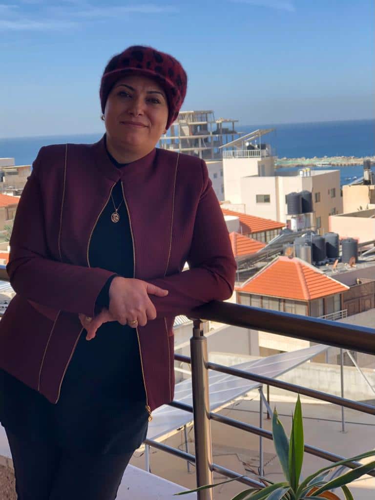 A woman in a burgundy jacket and beret cap posing for a photo on a balcony with the view of the city and sea in the background
