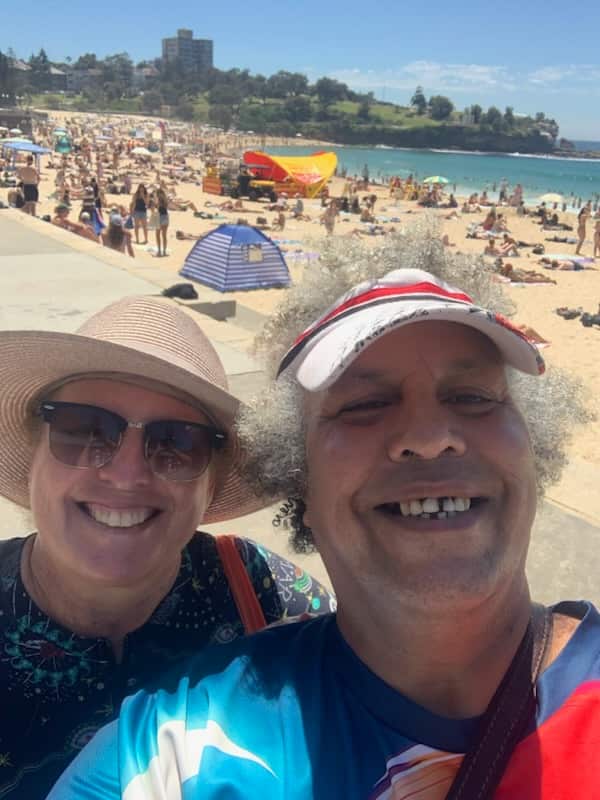 A smiling couple takes a selfie on a sunny day at a crowded beach with a lifeguard station and buildings in the background.