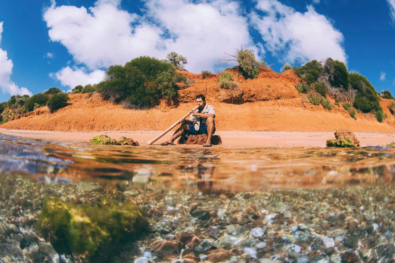 A man sitting on she shore, playing a didgeridoo.