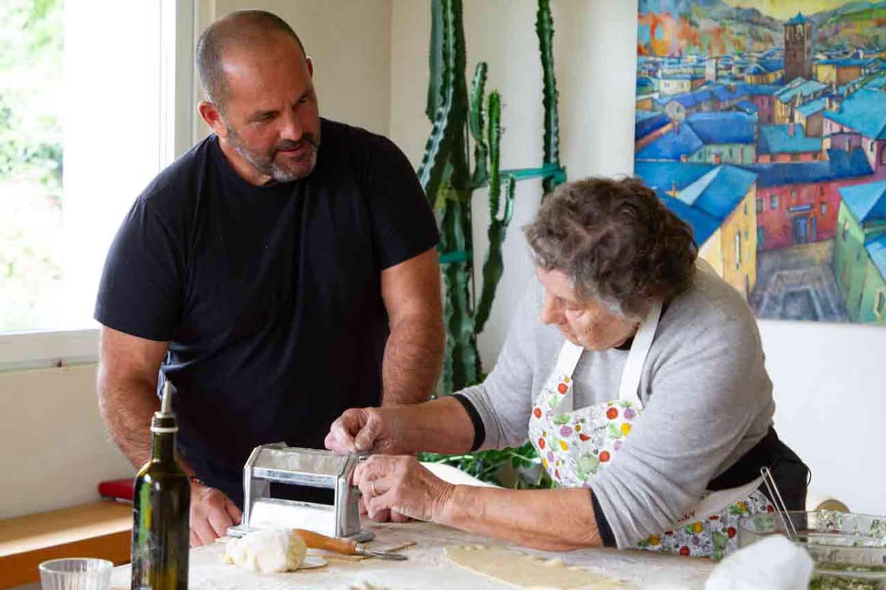 A man in a black t-shirt stands in a home kitchen beside an older woman in an apron, who is working a pasta rolling machine. 