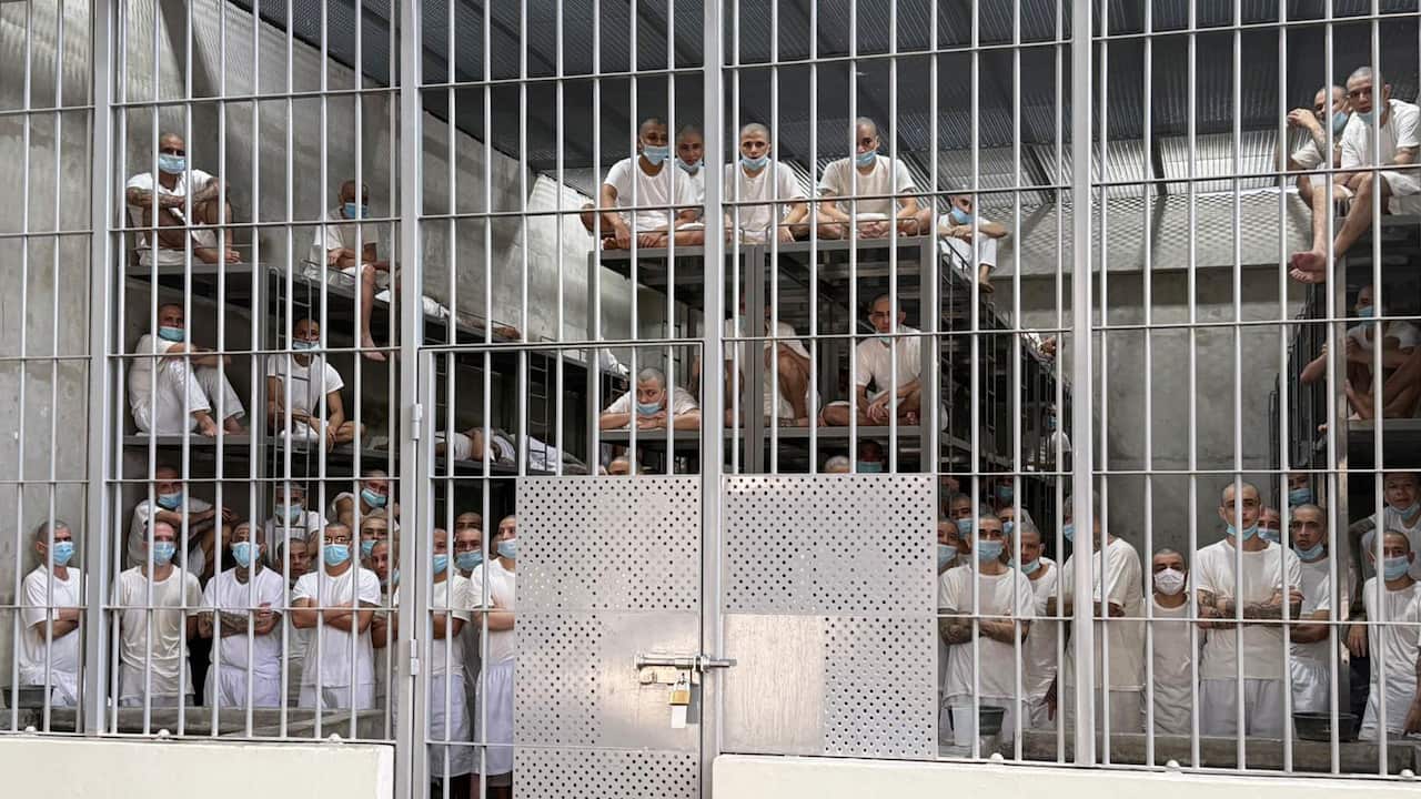 A group of men in white clothes and medical masks standing behind bars in a prison cell. Some are sitting on metal frames used as sleeping surfaces
