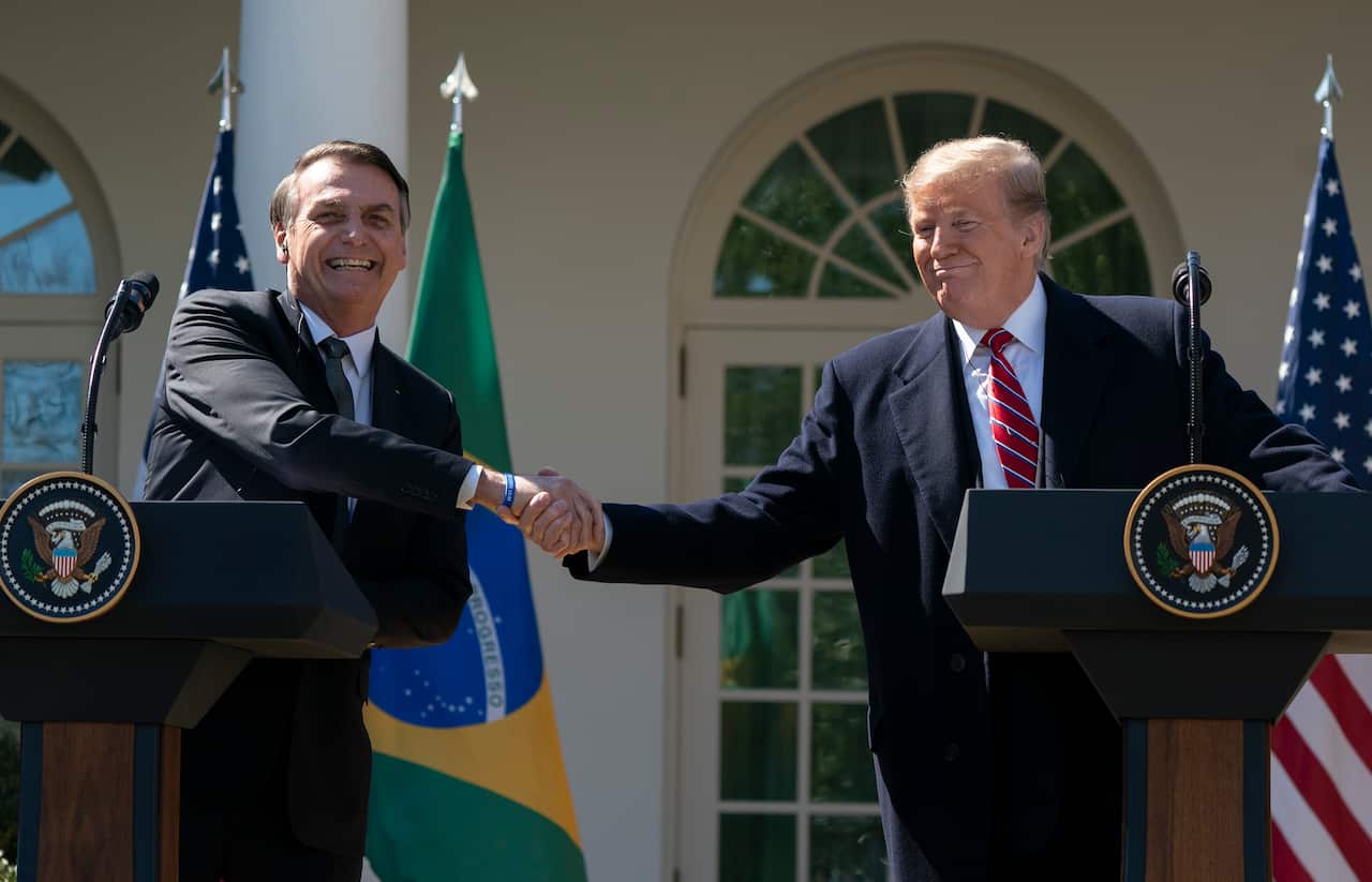 Jair Bolsonaro and Donald Trump shaking hands while standing at separate podiums outside the White House. The Brazilian and American flags are visible.