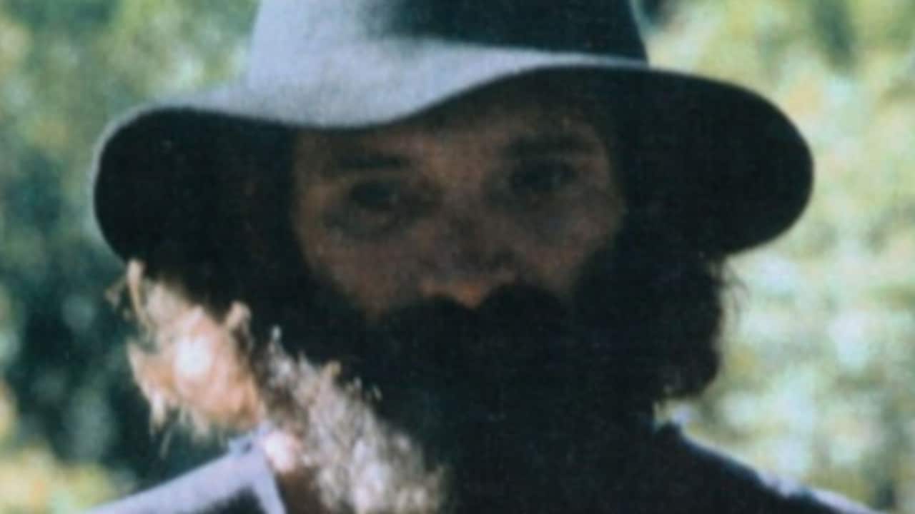 a close up shot of a man in a black felt hat with a long beard standing in front of forestry
