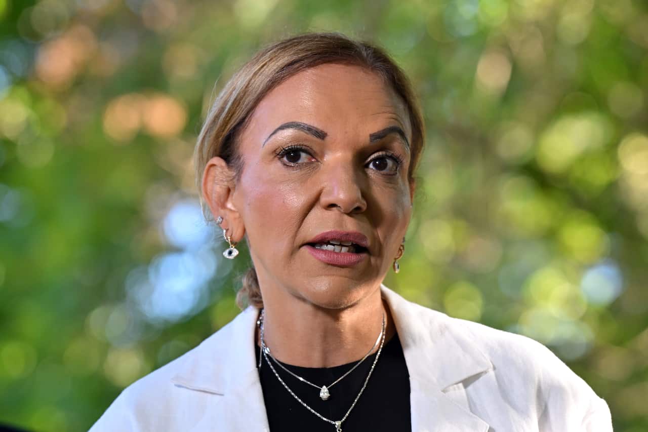 Anne Aly wearing a black shirt and white overshirt, standing underneath green trees.