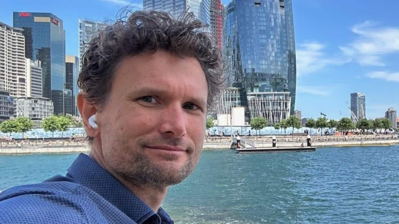 a man with curly hair and in a blue shirt taking a selfie in front of the water at Barangaroo