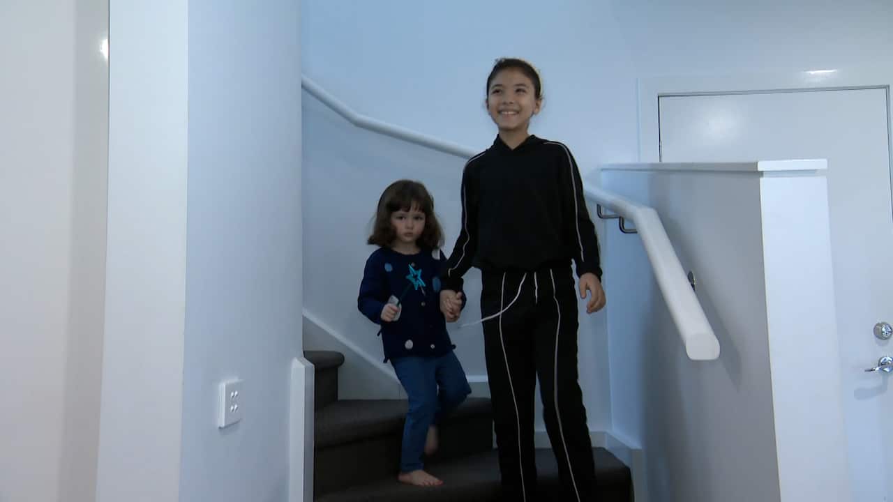 Two young girls come down a staircase covered in brown carpet, with a white handrail and white walls on either side of them.