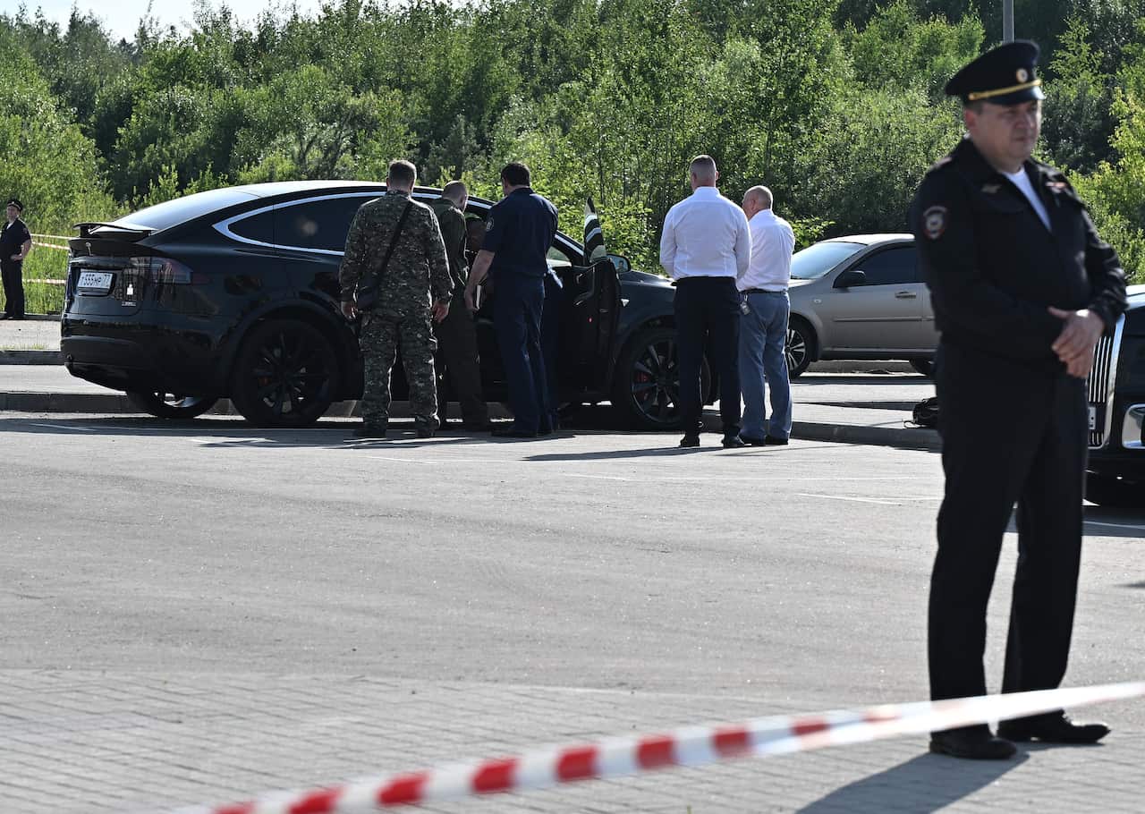 People standing near a black car outside, peering into it. A security officer is standing behind a line of tape.