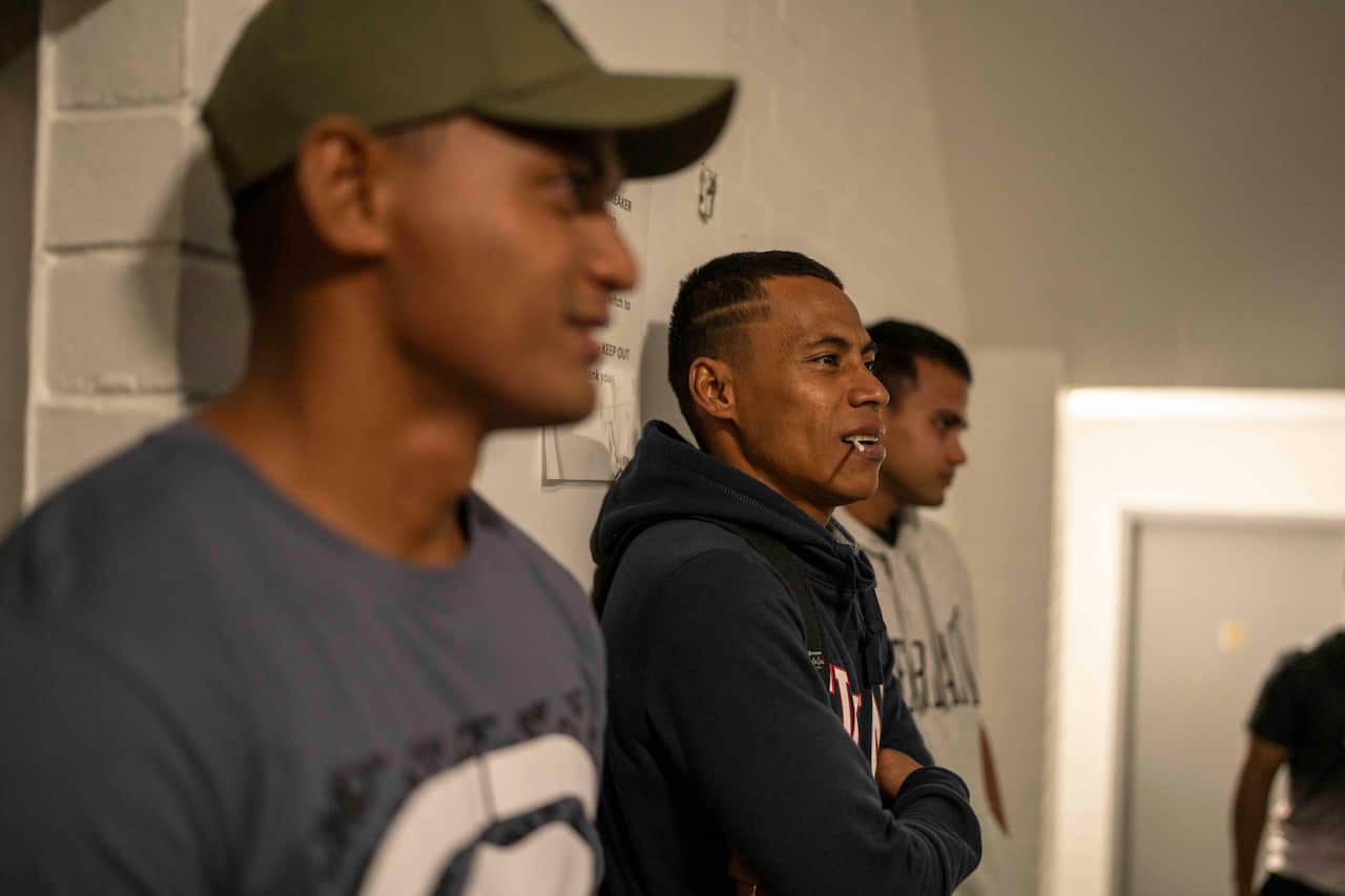 Three Pasifika men stand leaning against a wall outside a community hall, wearing casual clothing.