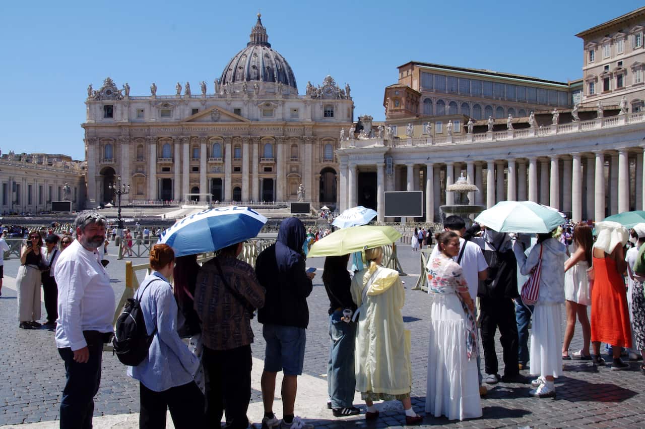 A crowd of tourists holding umbrellas stands in a sunlit plaza in front of St. Peter's Basilica under a clear blue sky.