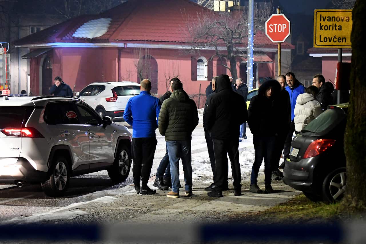 People standing near a red building, with snow on the ground and cars stopped.