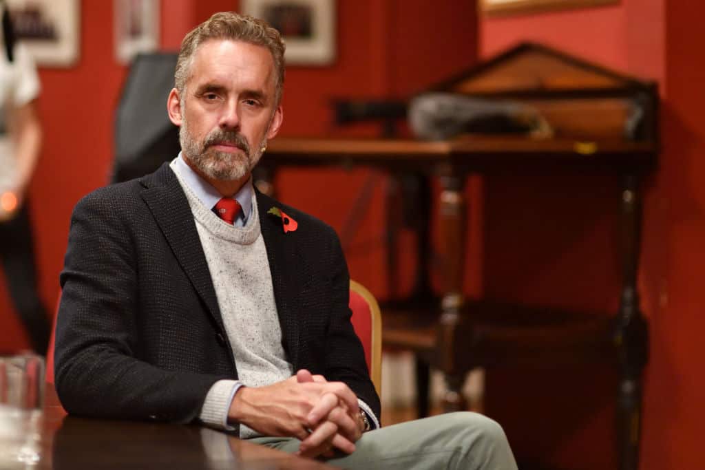 Jordan Peterson sits in a chair looking serious in a suit jacket. 