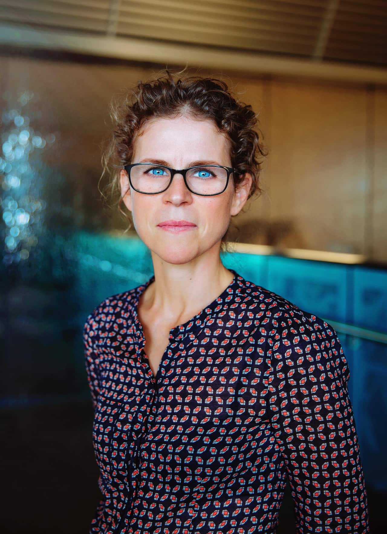 A headshot of a woman with curly brown hair, blue eyes, and black-rimmed glasses, wearing a dark patterned button-up shirt against a blurred, modern background.