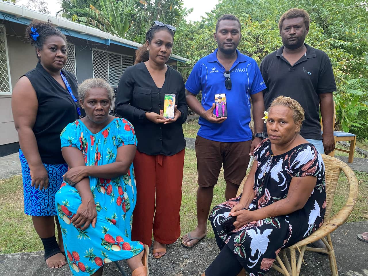 Two men and two women standing outside. Two women are sitting on chairs in front of them.