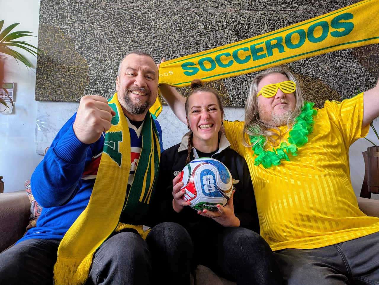 Two men and a woman dressed in green and gold and holding a Socceroos scarf and a football on a couch