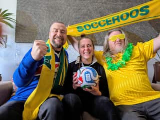 Two men and a woman dressed in green and gold and holding a Socceroos scarf and a football on a couch