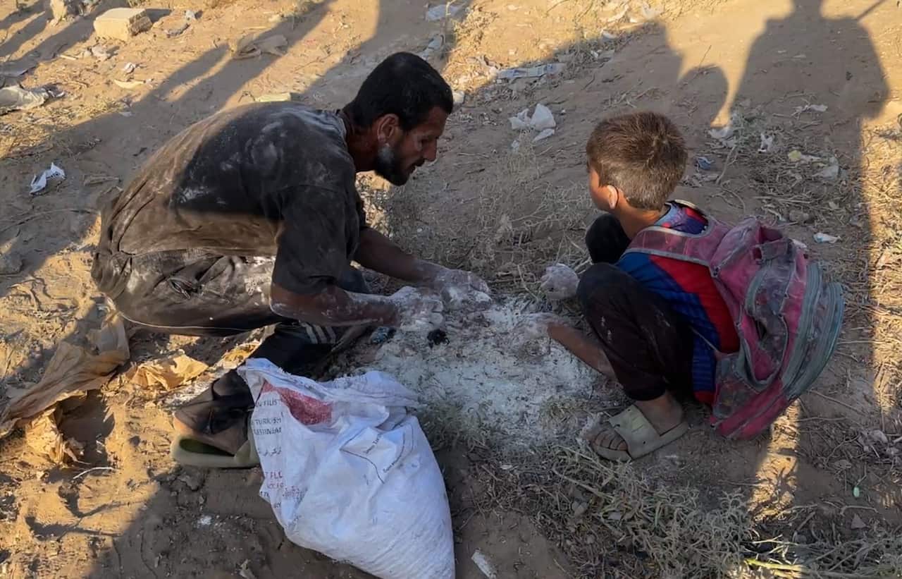 A man and child kneel to pick flour out of the dirt.