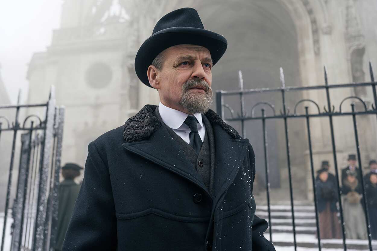 Man in dark coat and hat stands in front of metal fence, gothic building behind him. 