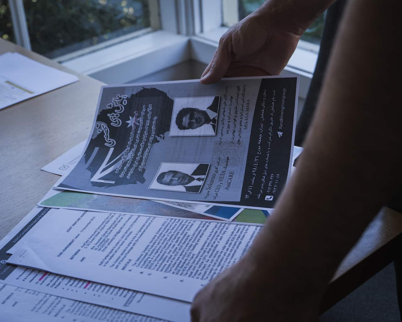 A person's hands are holding up a flyer with two headshots and text in both English and what appears to be Persian. The flyer is part of a stack of documents, including a joint venture agreement, laid out on a table near a window