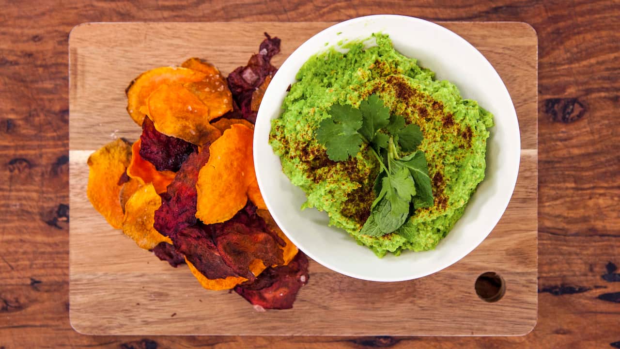 Seen from overhead, a white bowl filled with a green dip sits on a wooden board, alongside a pile of vegetable crisps. 