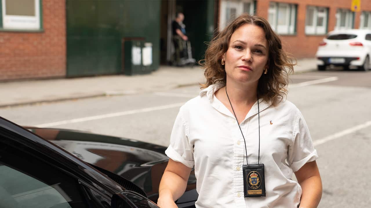 A woman wearing a short-sleeved white shirt stands beside a black car on a street, with a police badge hanging from a lanyard around her neck. Brick buildings and parked cars are visible in the background