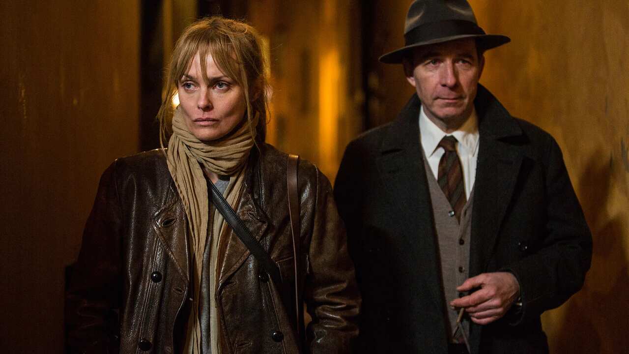 A woman in an leather jacket and a man wearing a suit and hat walk down a dark street