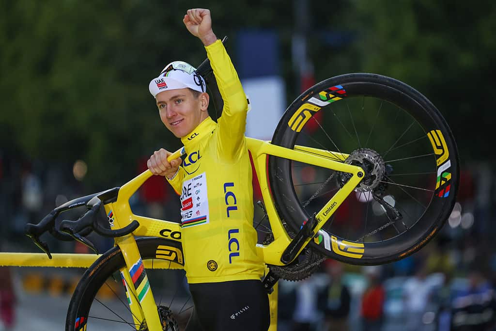 A man wearing yellow cycling jersey and a white cap holds his left arm up in triumph as he carries a yellow bicycle over his right shoulder